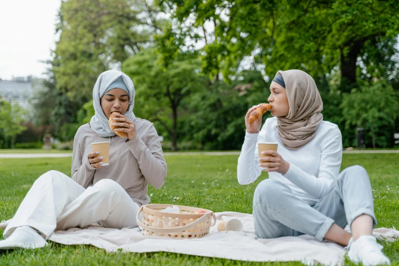 a couple of women sitting on top of a grass covered field, a picture, shutterstock, hurufiyya, eating garlic bread, muslim, drinking a strawberry iced latte, sydney park