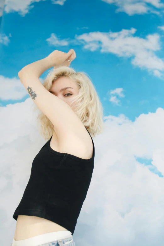 a woman standing in front of a mural of clouds, an album cover, unsplash, photorealism, pale skin curly blond hair, with arms up, charli bowater, she is wearing a black tank top