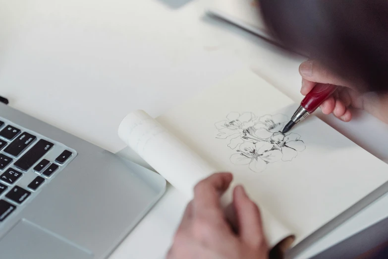 a person drawing on a piece of paper next to a laptop, inspired by James Jean, trending on pexels, arbeitsrat für kunst, floral design, shawn coss, tattoo design, lithography