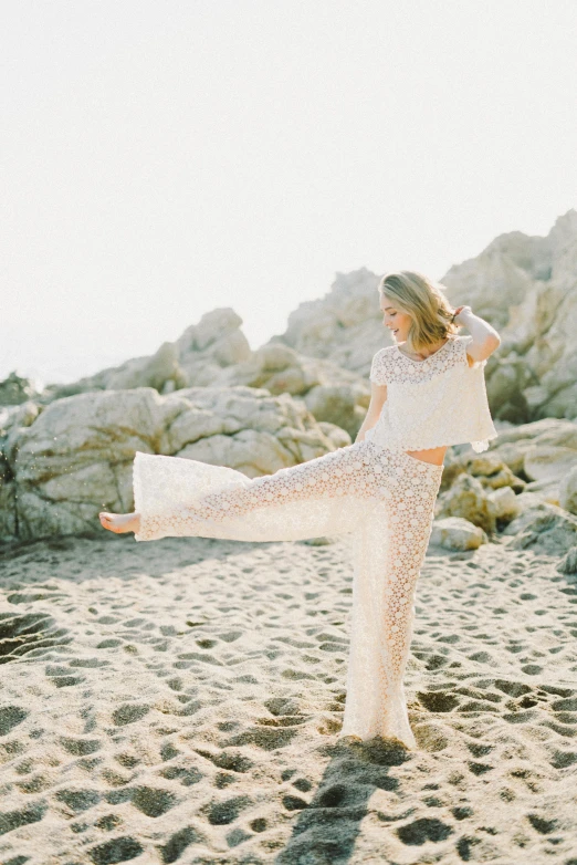 a woman standing on top of a sandy beach, by Robbie Trevino, arabesque, in a white boho style studio, standing on rocky ground, sparkling, transparencies