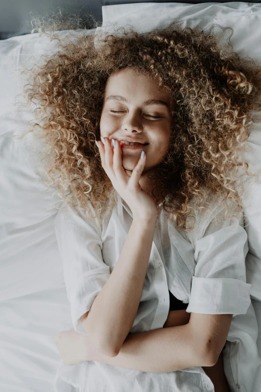 a woman laying in bed with her eyes closed, trending on pexels, renaissance, curly middle part haircut, huge smile, teenager girl, hand on her chin