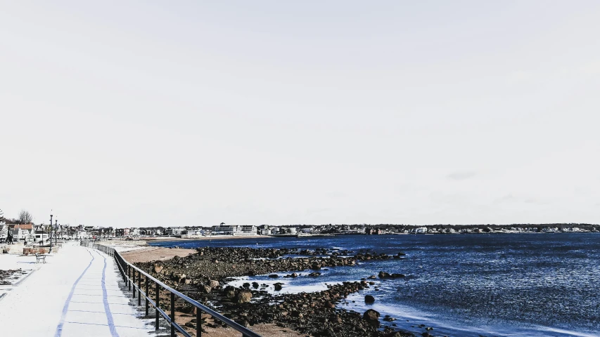 a bench sitting on the side of a road next to a body of water, unsplash, happening, rocky beach, wood pier and houses, distant cityscape, photographic print