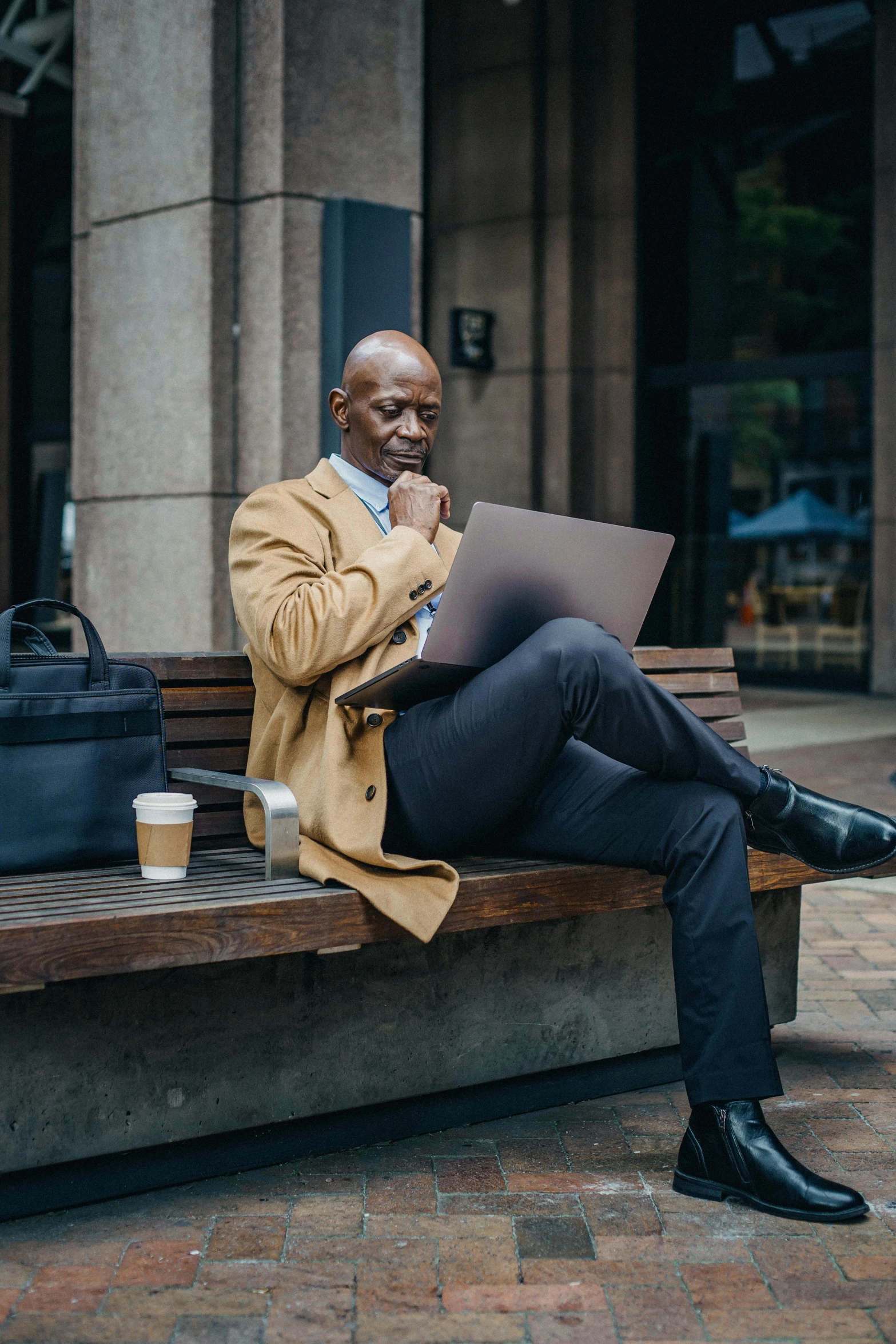 a man sitting on a bench with a laptop, trench coat and suit, black man, advanced economy, bald man