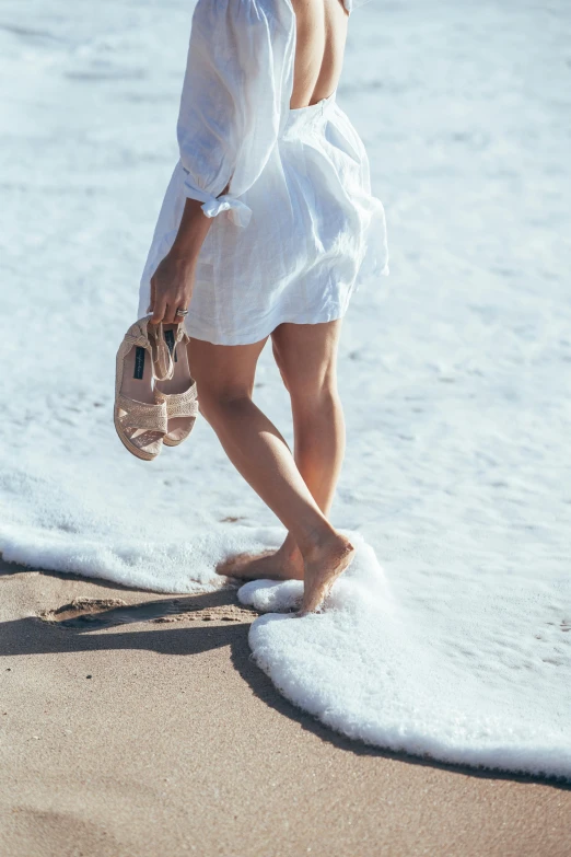 a woman walking on a beach next to the ocean, slippers, white water, light tan, battered