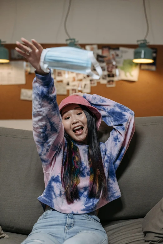 a girl sitting on a couch with her hands in the air, a picture, inspired by Kim Jeong-hui, trending on pexels, with rap cap on head, soey milk, tie-dye, wearing a blue hoodie
