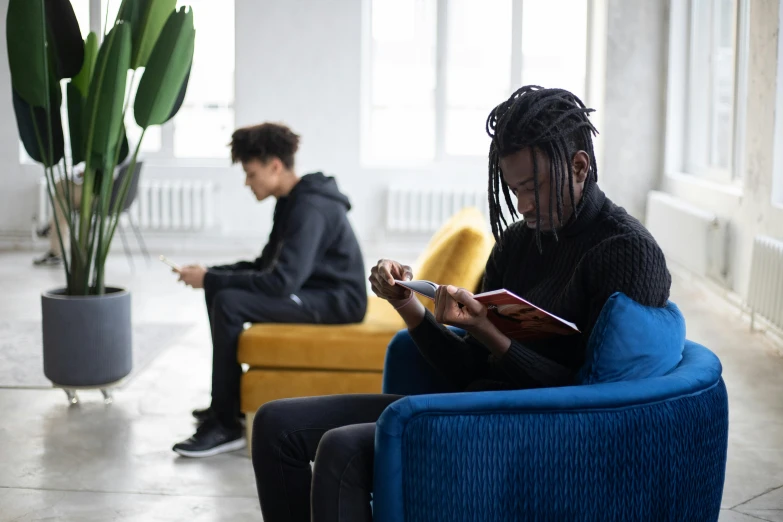 a man sitting in a chair reading a book, pexels contest winner, two young men, black teenage boy, in a open-space working space, focus on two androids