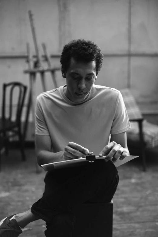 a black and white photo of a man sitting on a stool, inspired by Max Klinger, hyperrealism, writing on a clipboard, robert sheehan, square, adam sandler