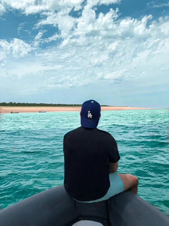 a man sitting on top of a boat in the ocean, wearing a baseball hat, trending photo, australian beach, distance view