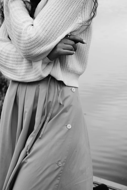 a woman standing next to a body of water, a black and white photo, unsplash, renaissance, white shirt and grey skirt, wearing a white sweater, close up details, folds