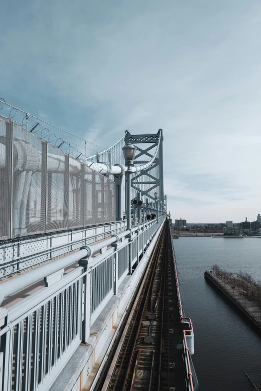 a train traveling over a bridge next to a body of water, by Jacob Burck, pexels contest winner, steel archways, looking over city, new jersey, grey