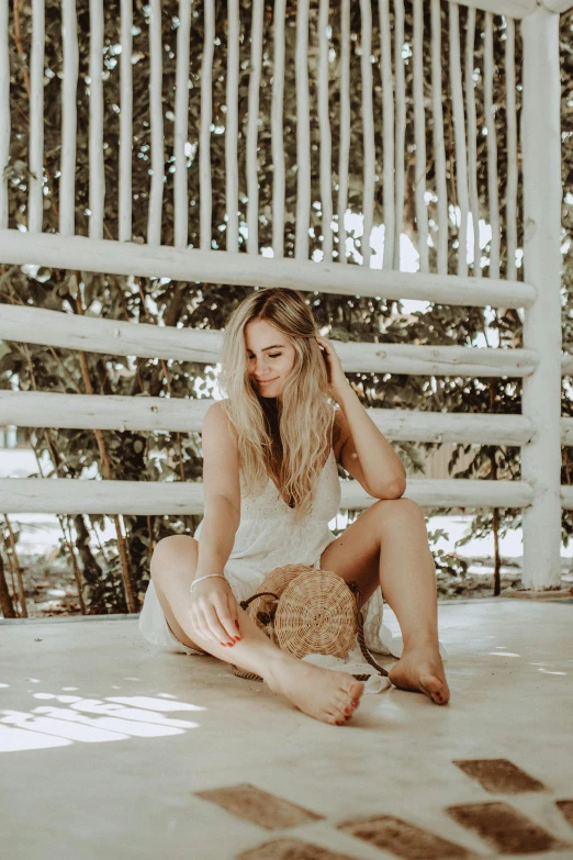 a woman sitting on the ground in front of a white fence, bali, tan skin, with long blond hair, item