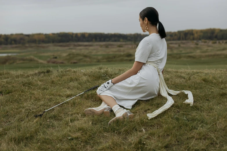 a woman sitting on top of a grass covered field, holding a staff, wearing off - white style, louise zhang, hammershøi