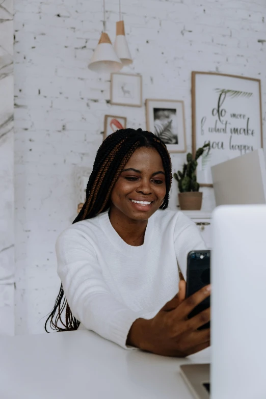 a woman sitting at a desk using a cell phone, a picture, trending on pexels, happening, wearing a white sweater, black woman, welcoming grin, using a macbook