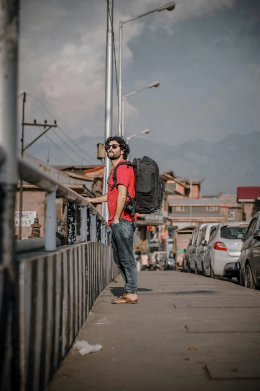 a man with a backpack standing on a sidewalk, inspired by Steve McCurry, pexels contest winner, happening, mountains in the background, avatar image, standing in township street, model pose