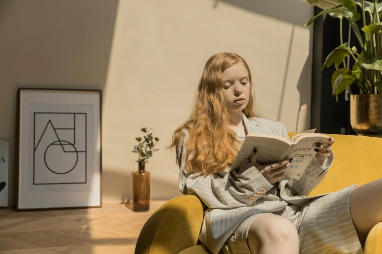 a woman sitting on a yellow couch reading a book, inspired by Balthus, pexels contest winner, long ginger hair, sunny morning light, wearing a grey robe, gif