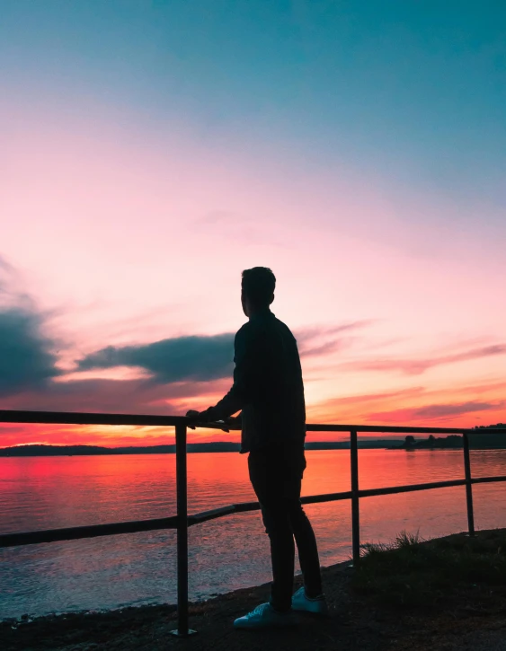 a man standing on top of a pier next to a body of water, pexels contest winner, orange / pink sky, profile picture, lgbtq, outlined silhouettes