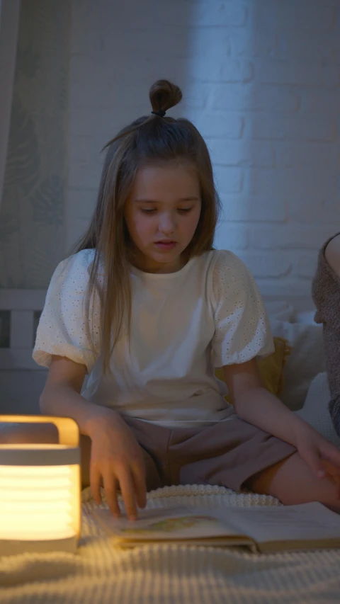 a little girl sitting on a bed reading a book, inspired by Henry Ossawa Tanner, pexels, realism, connected with glowing tubes 8 k, gif, himalayan rocksalt lamp, film screenshot