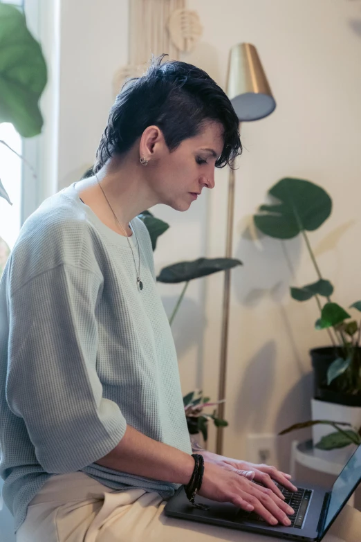 a woman sitting on a bed using a laptop computer, a portrait, inspired by Elsa Bleda, trending on unsplash, short green bobcut, wearing a light blue shirt, standing on a desk, botanicals