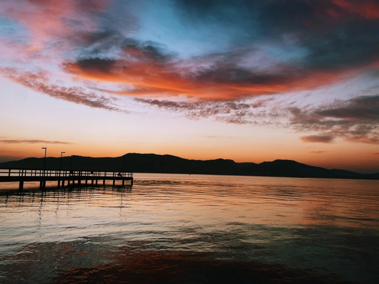 a pier sitting on top of a body of water, pexels contest winner, mountains and colorful sunset!!, instagram post, sunset with cloudy skies, slightly tanned