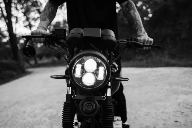 a man riding a motorcycle down a dirt road, a black and white photo, by Kristian Zahrtmann, custom headlights, close up high detailed, cafe racer, multiple lights