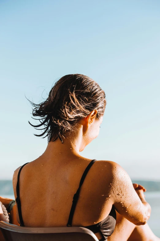 a woman sitting in a chair on the beach, unsplash, showing her shoulder from back, with textured hair and skin, sun glare, wet hair