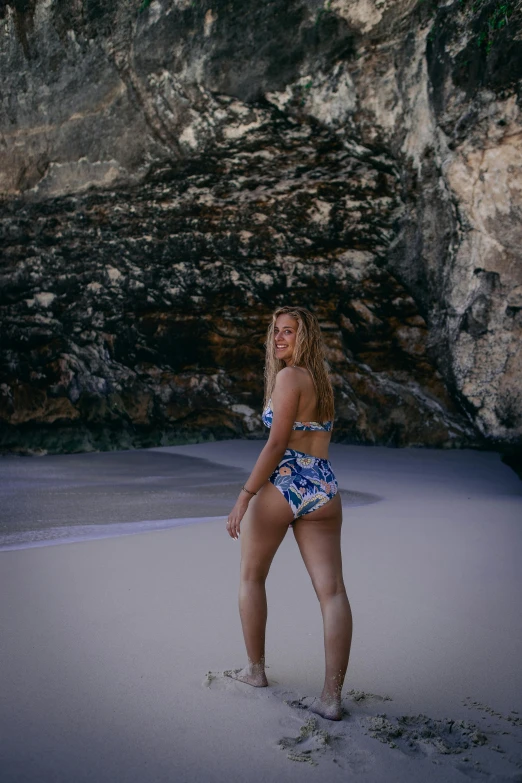 a woman in a bikini standing on a beach, a picture, by Lee Loughridge, unsplash, renaissance, standing on rocky ground, low quality photo, back turned, floral
