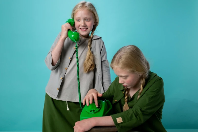 two young girls are talking on the telephone, inspired by Elsa Beskow, wearing green jacket, official product photo, backdrop, girl wearing uniform