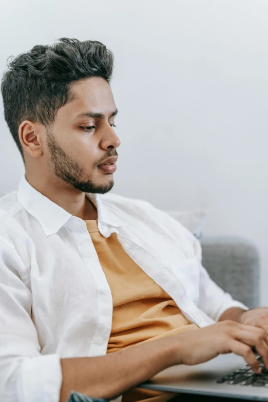 a man sitting on a couch using a laptop, trending on pexels, renaissance, wearing a white button up shirt, indian, looking serious, non binary model