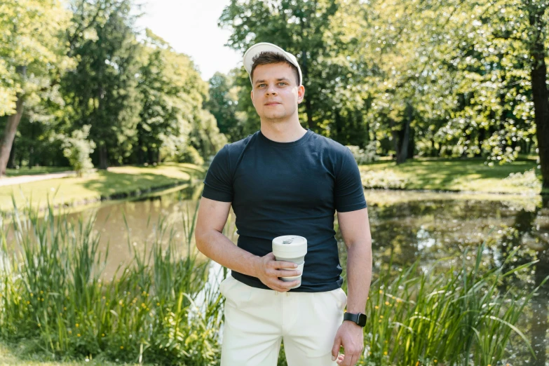 a man standing next to a lake holding a cup of coffee, a portrait, by Adam Marczyński, wearing golf shorts, avatar image, max verstappen, in a park