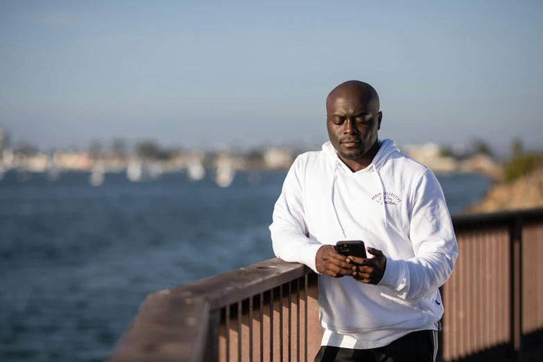 a man standing on a bridge looking at his cell phone, a portrait, by Stokely Webster, happening, wearing a track suit, long beach background, avatar image, portrait of bald