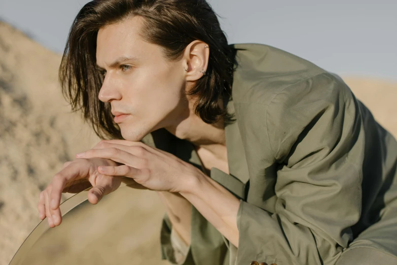 a close up of a person holding a mirror, an album cover, trending on pexels, bauhaus, attractive man, brown colored long hair, green and brown clothes, on the desert