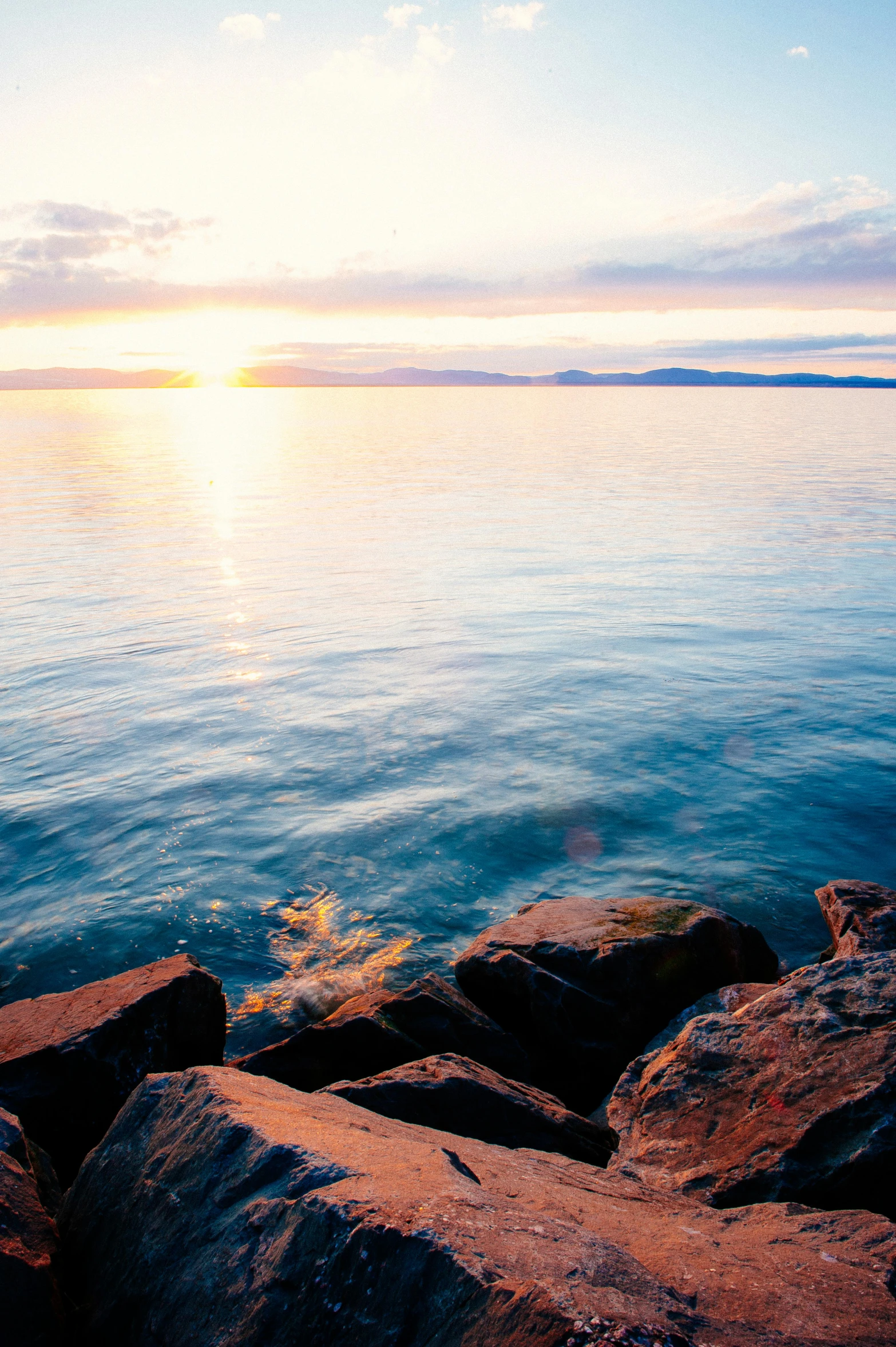 a sunset over a body of water with rocks in the foreground, midday sunlight, superior look, lake foreground, shoreline