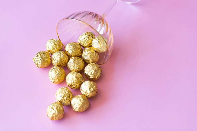 a glass filled with gold foiled chocolates on a pink surface, a still life, by Julia Pishtar, pexels, some spherical, slightly tanned, tribbles, white gold