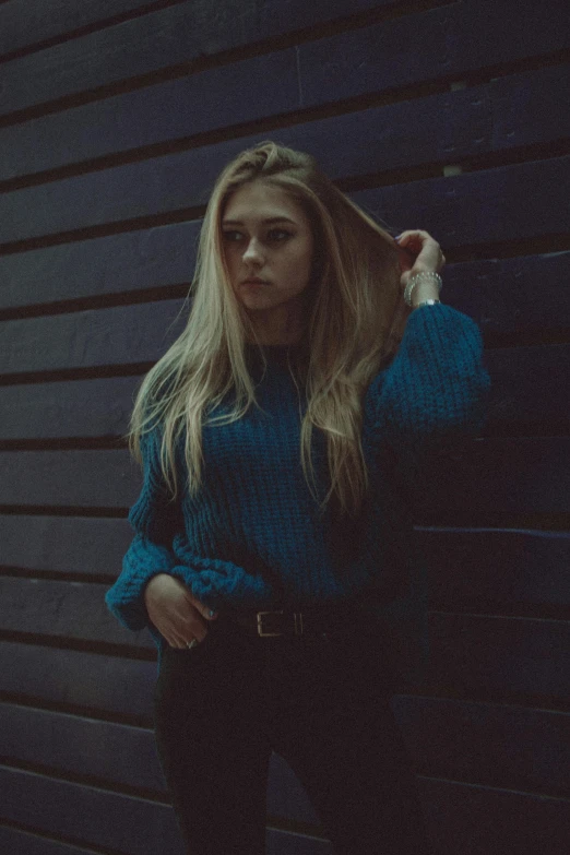 a woman in a blue sweater leaning against a wall, inspired by Elsa Bleda, unsplash, aestheticism, ava max, standing with a black background, grainy quality, gif