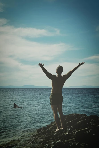 a man standing on top of a rock next to the ocean, hands in the air, shark man, colour photo, summer feeling