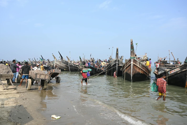 a group of people standing on top of a beach next to a body of water, hurufiyya, fishing boats, ranjit ghosh, maintenance photo