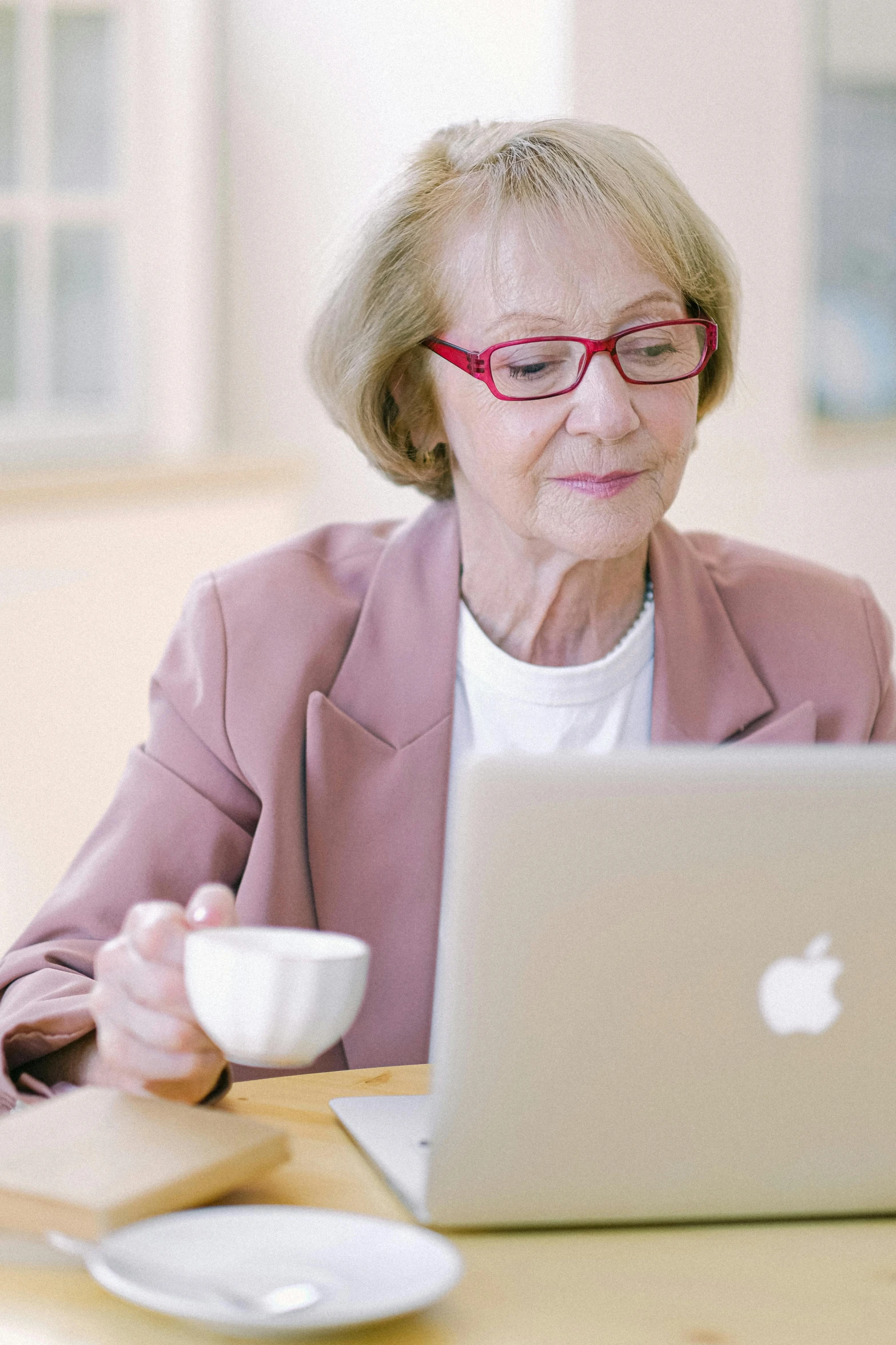 a woman sitting at a table with a laptop and a cup of coffee, by Jan Cox, with apple, an elderly, intelligent, music video