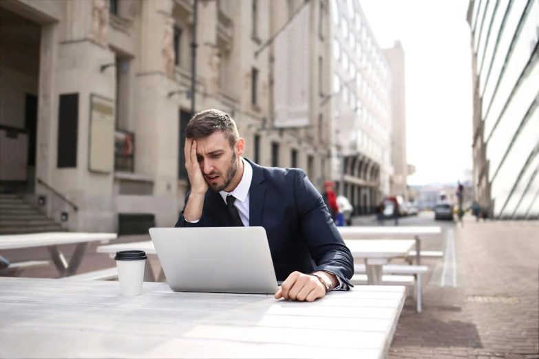 a man in a suit sitting at a table with a laptop, by Julia Pishtar, pexels, on a hot australian day, people panicking, street - level, denys tsiperko