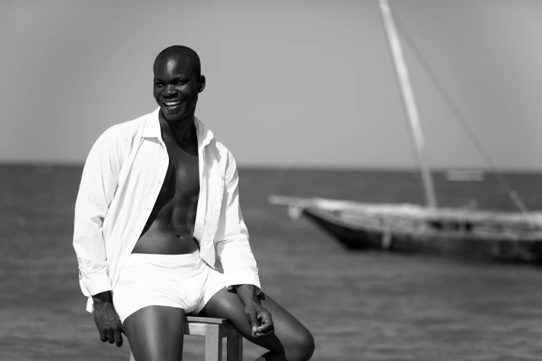 a black and white photo of a man sitting on a chair, by Maurycy Gottlieb, adut akech, posing on the beach, wearing a white button up shirt, on a yacht at sea