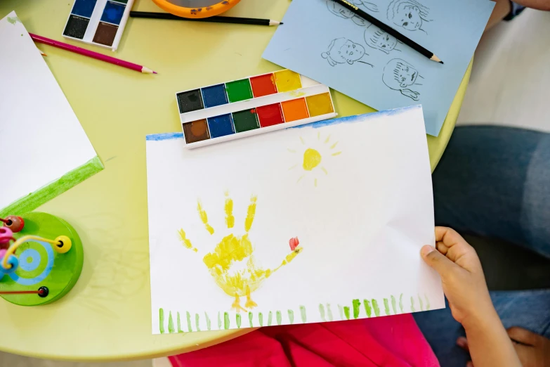 a person sitting at a table holding a piece of paper, a child's drawing, inspired by Helen Frankenthaler, pexels contest winner, 🎨🖌, finger painting, sunny day, getty images