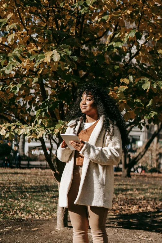 a woman standing under a tree holding a cup of coffee, an album cover, trending on pexels, happening, using a magical tablet, light brown coat, black young woman, professional iphone photo