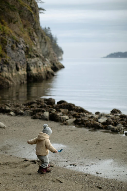 a small child standing on a beach next to a body of water, british columbia, slide show, explore, a cozy