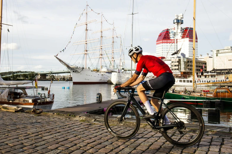 a man riding a bike next to a body of water, by Tom Wänerstrand, pexels contest winner, wearing a red captain's uniform, harbour in background, kirsi salonen, nike cycling suit