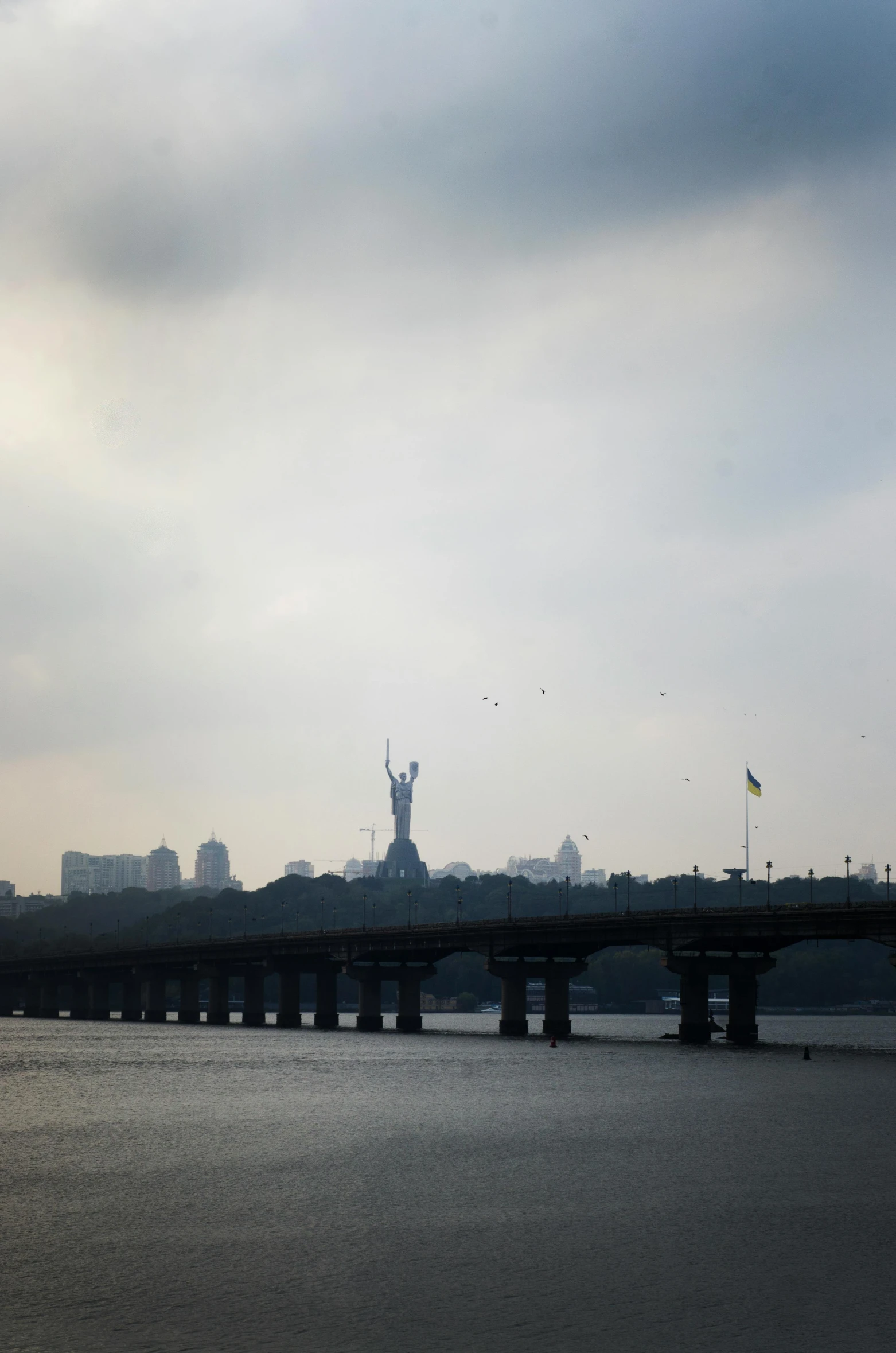 a bridge over a body of water under a cloudy sky, a statue, neo kyiv, silhouette :7, very hazy, photographed for reuters