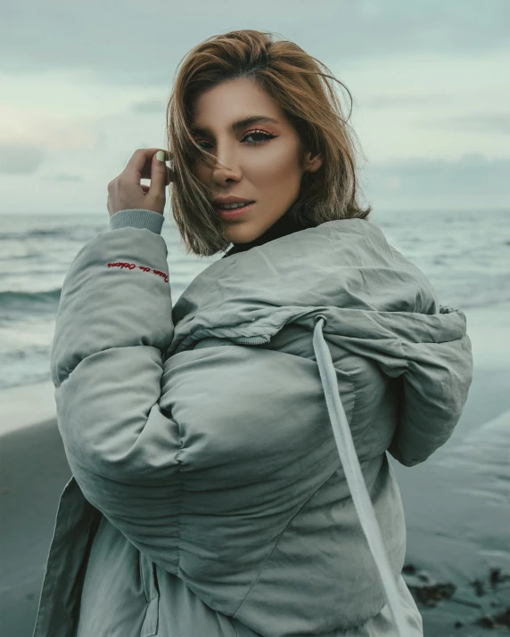 a woman standing on top of a beach next to the ocean, an album cover, inspired by Elsa Bleda, trending on pexels, graffiti, model wears a puffer jacket, grey rubber undersuit, young beautiful amouranth, tight wrinkled cloath