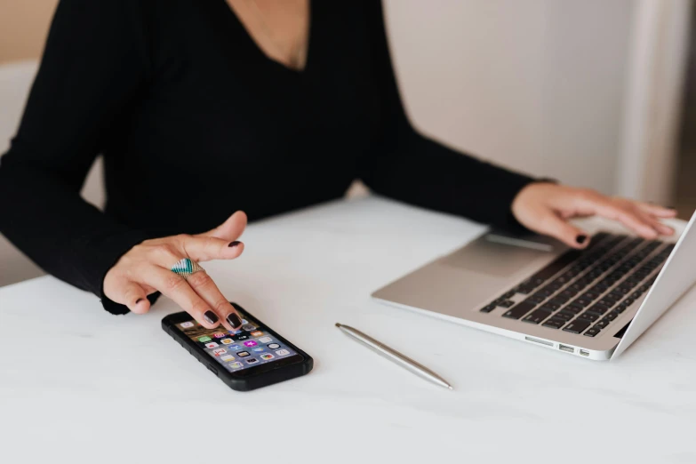 a woman sitting at a table with a laptop and a cell phone, trending on pexels, corporate phone app icon, black hoodie techie, hovering indecision, 1 4 5 0