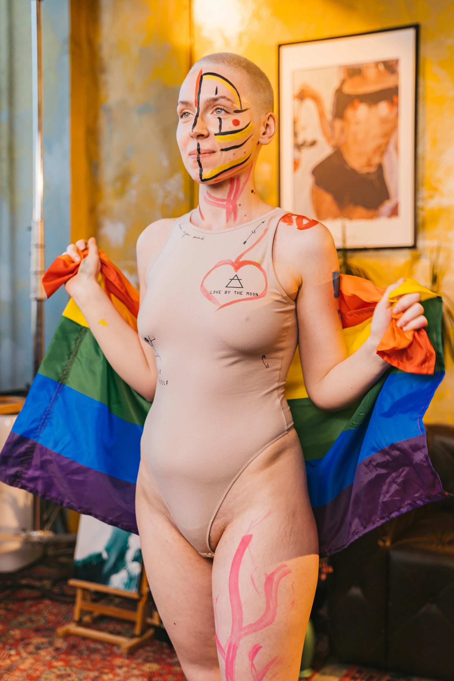 a woman standing in a living room holding a rainbow flag, by Jessie Alexandra Dick, latex flesh and facial muscles, bodysuit, with a white complexion, cracked body full of scars