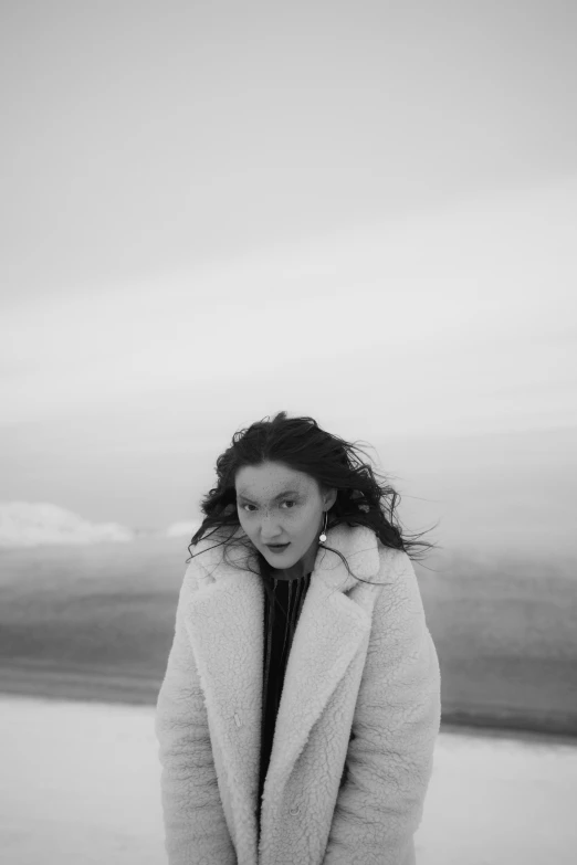 a woman standing in front of a body of water, a black and white photo, wearing a white winter coat, lorde, promotional image, windy beach