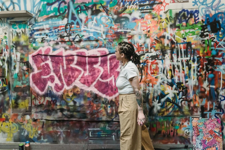 a woman standing in front of a wall covered in graffiti, trending on pexels, wearing cargo pants, tourist photo, queer woman, background image