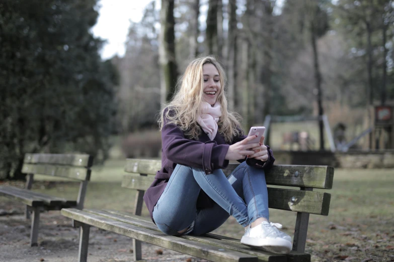 a woman sitting on top of a wooden bench, a picture, pexels, happening, she is holding a smartphone, a blond, teenager, a park